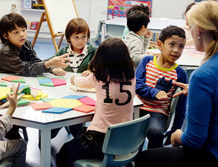 elementary school kids sitting around a table at school doing an art project with the teacher next to them
